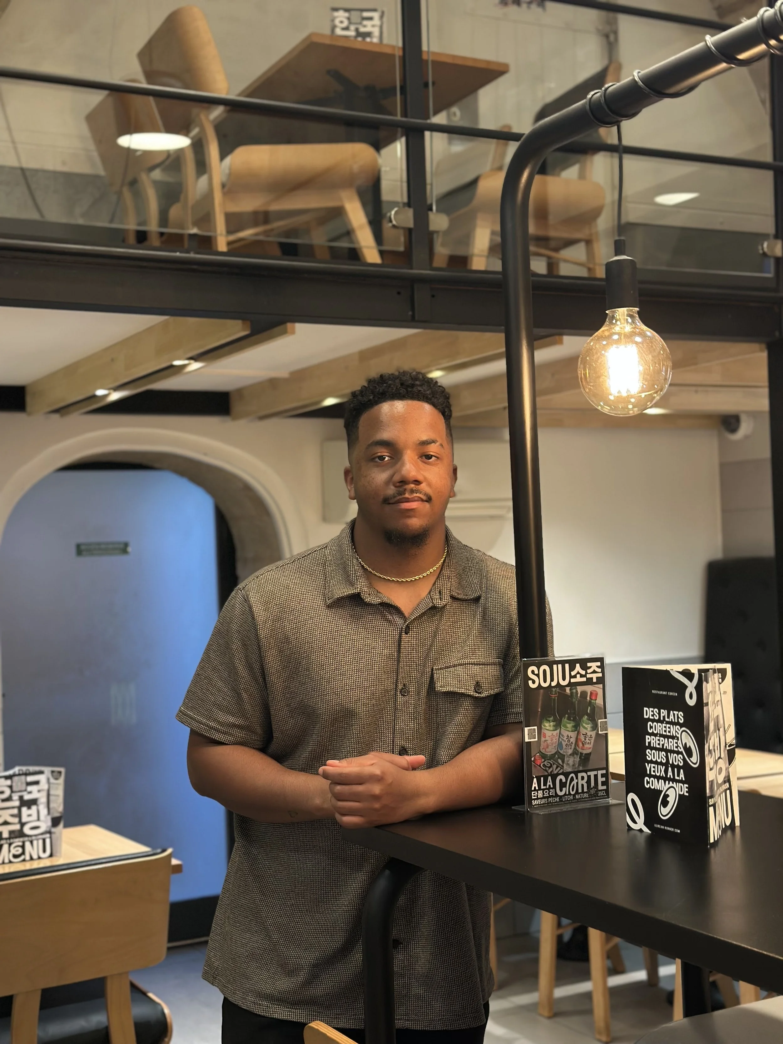 A young man standing inside a restaurant, wearing a short-sleeved collared shirt and a gold necklace, behind a black table with menus and a sign for Korean-style soy sauce marinated dishes.