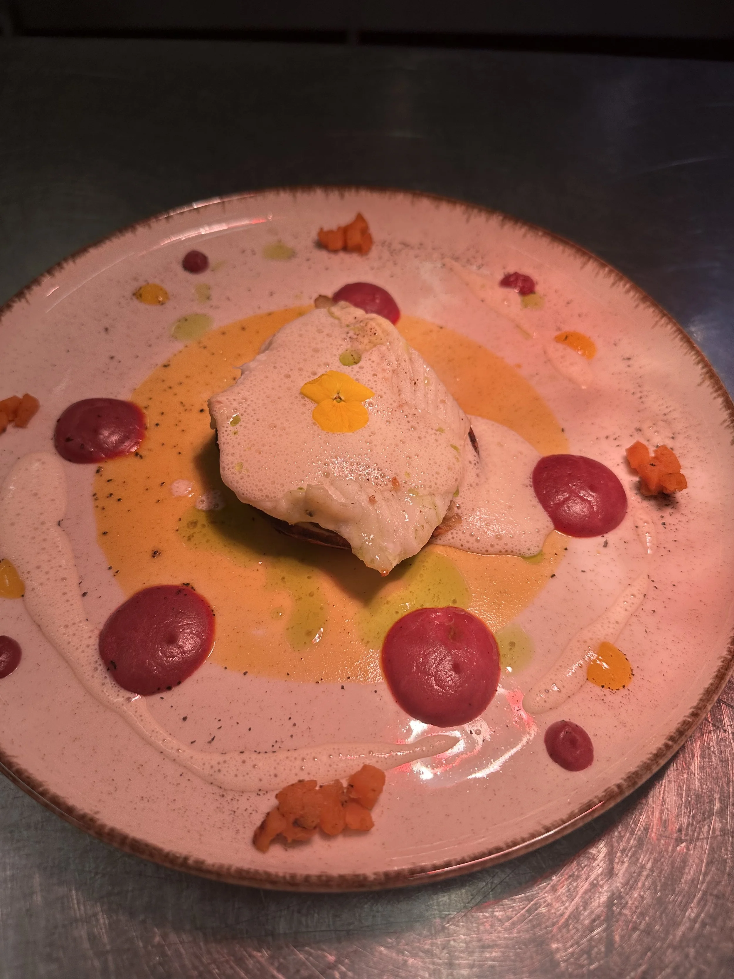 A plated gourmet dish featuring a piece of fish topped with foam, garnished with edible flowers and surrounded by various colorful dots and small bits of garnish.