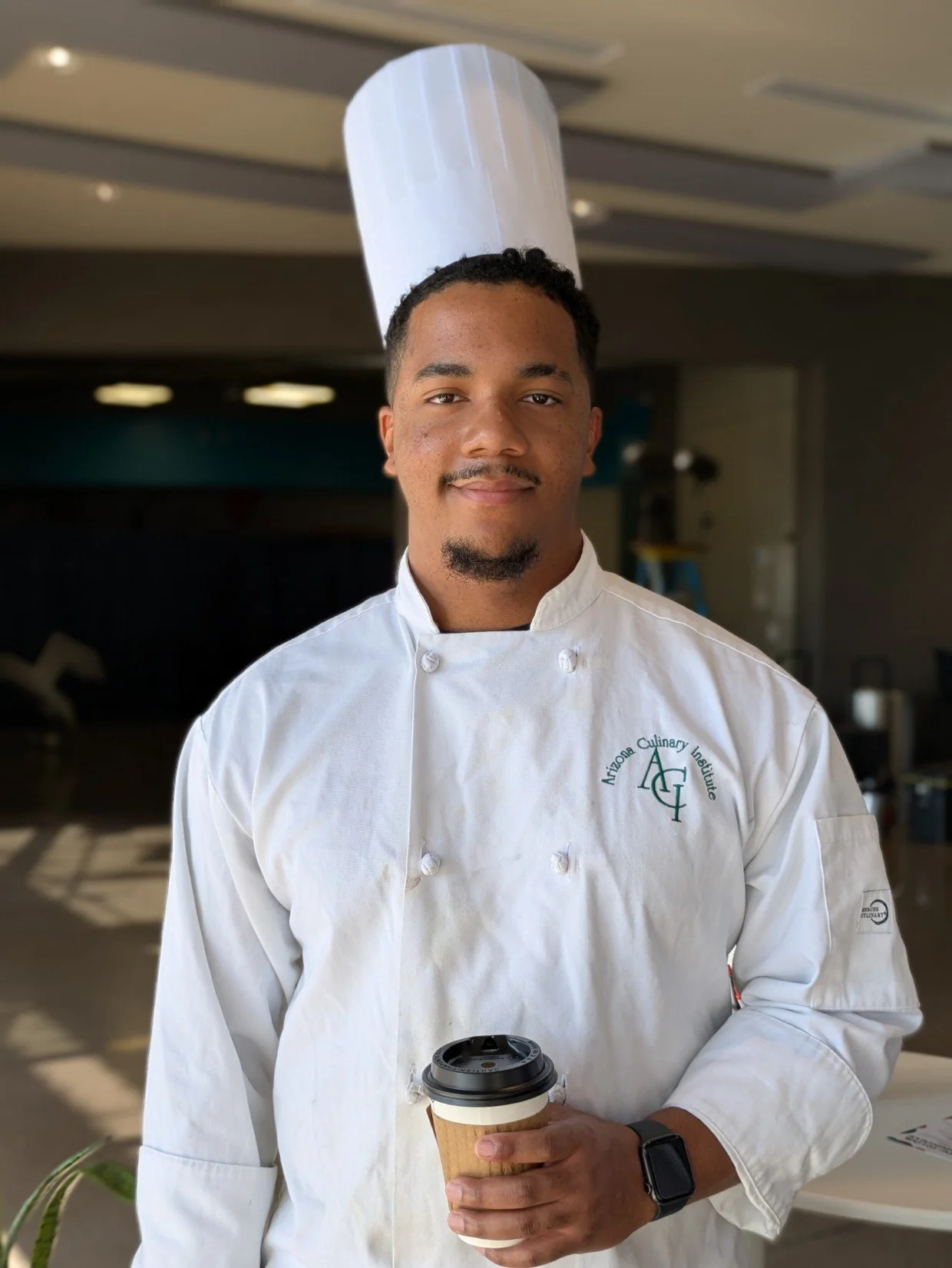 A male chef in a white chef's coat and a tall white chef's hat, holding a coffee cup, standing indoors in a kitchen or restaurant setting.