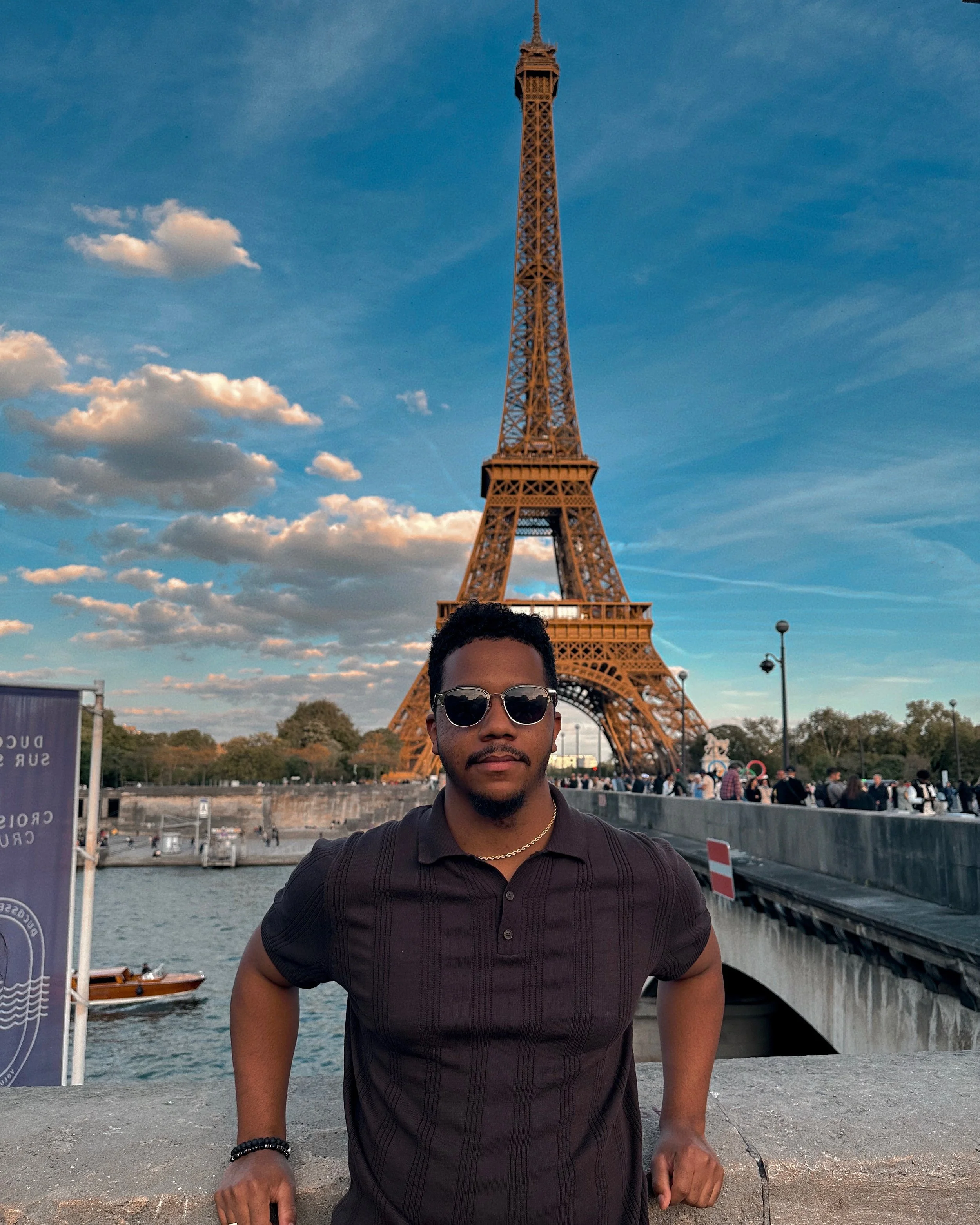 A man wearing sunglasses and a black polo shirt posing in front of the Eiffel Tower in Paris, France during the day with a partly cloudy sky.
