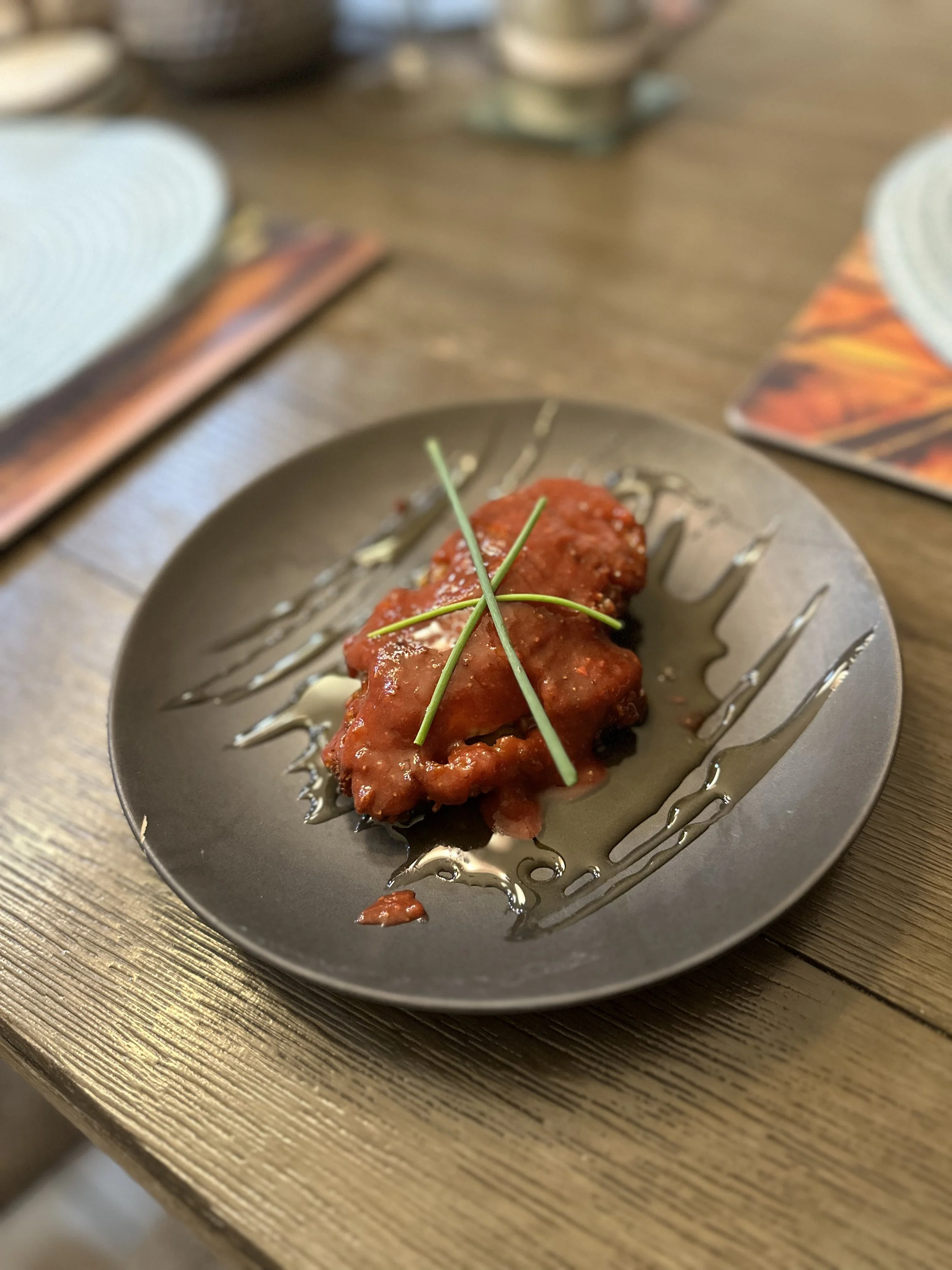 A plate with a piece of fried chicken covered in red sauce, garnished with green herbs, on a black plate with sauce drizzle, on a wooden table.