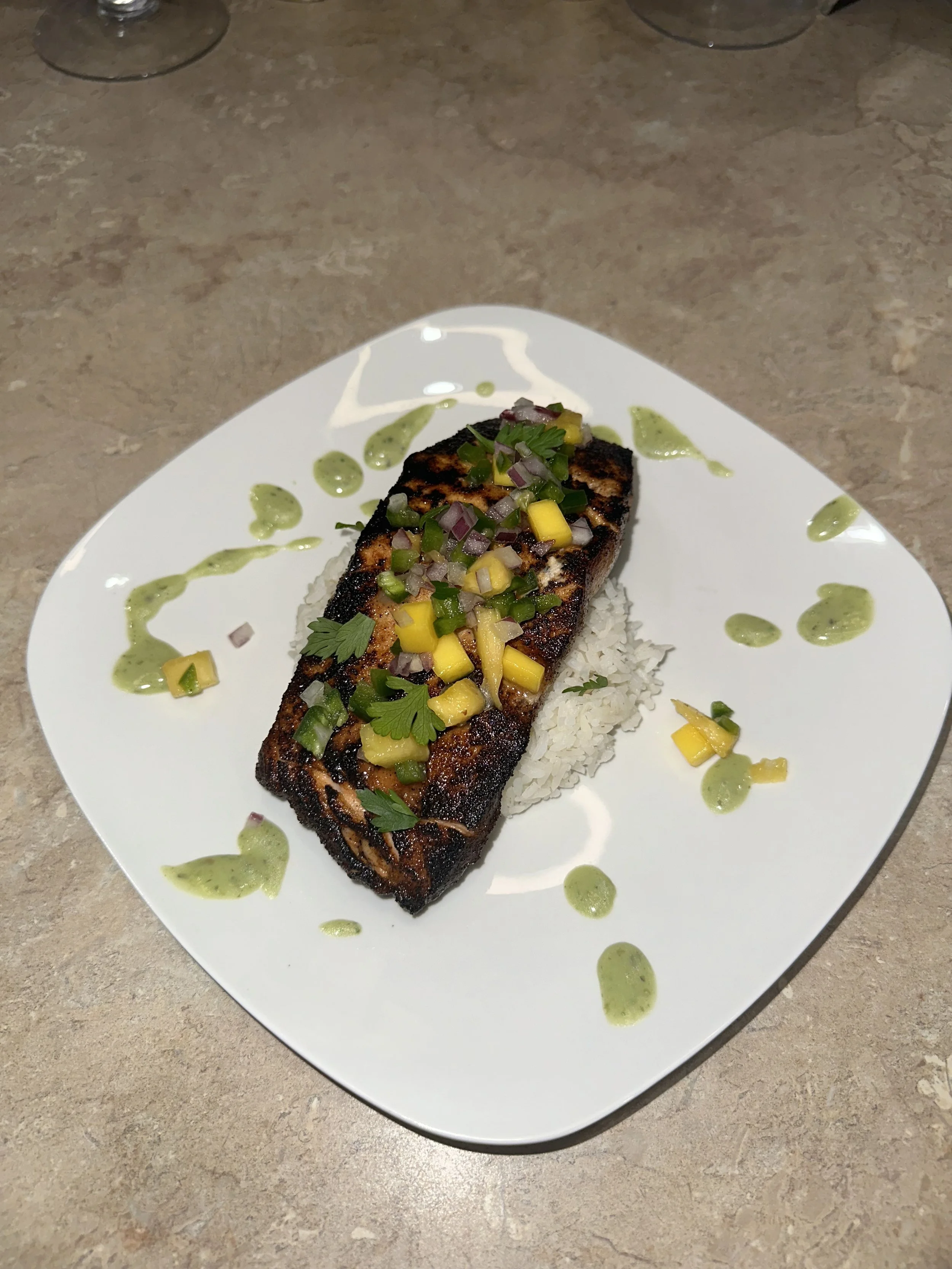 Grilled salmon fillet on rice, garnished with chopped mango, onion, cilantro, and green sauce, served on a white plate.