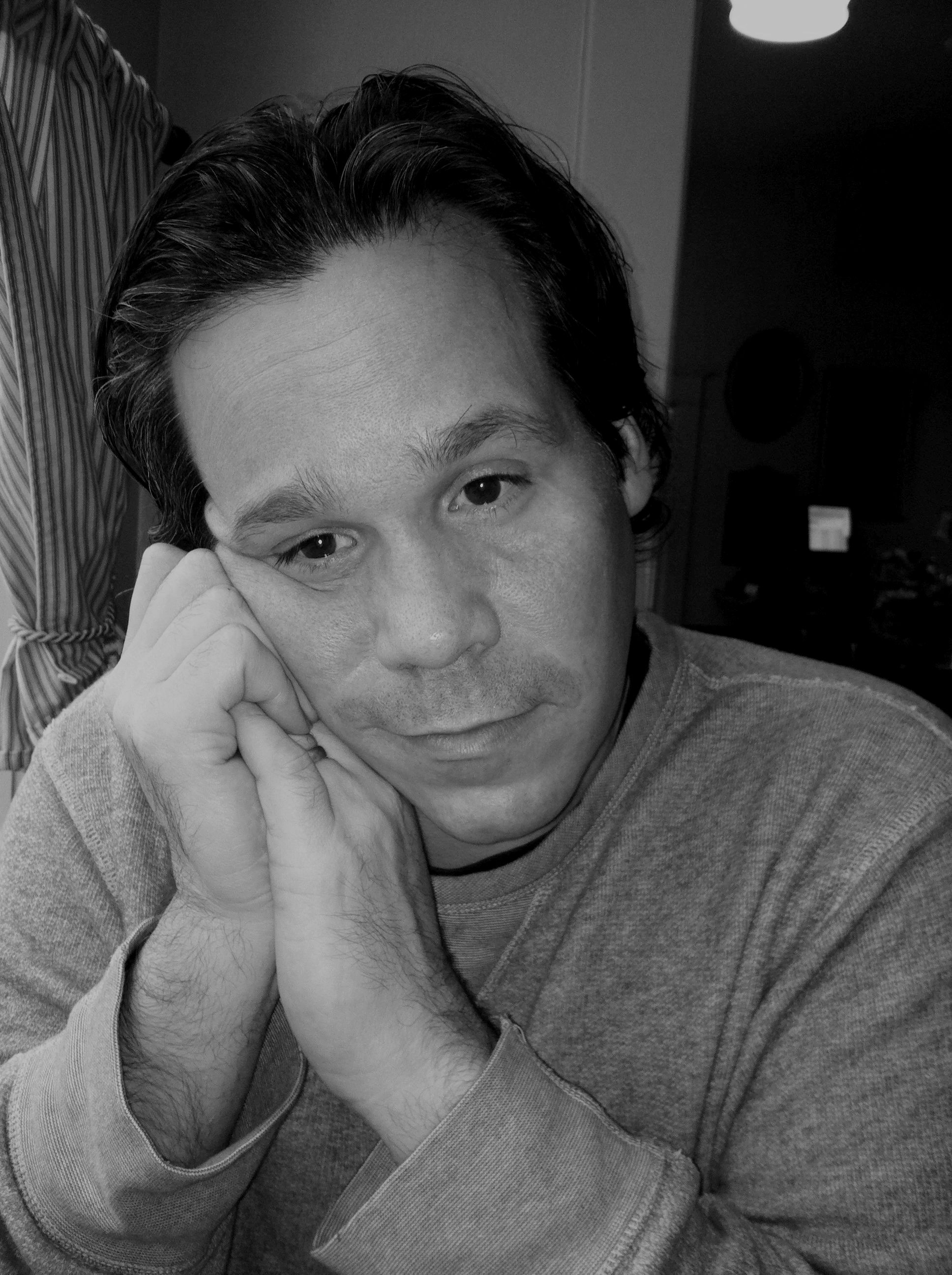 A man with dark, slicked-back hair resting his chin on his hand, looking directly at the camera, in a black and white photo.