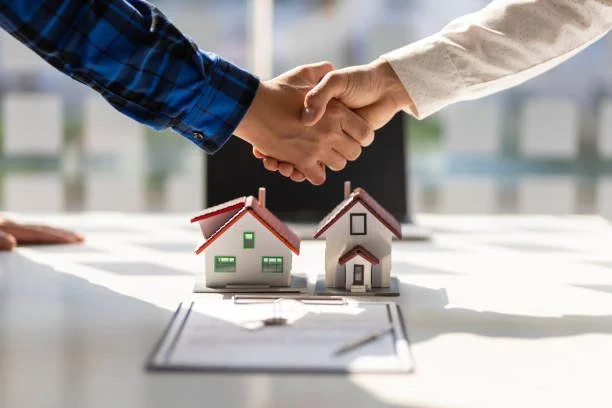 Two people shaking hands over a table with miniature house models and a clipboard in front of them, indicating a home agreement or real estate deal.