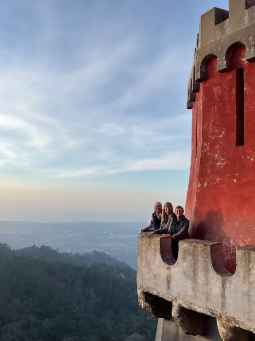 Three people sitting on a ledge of a castle tower, overlooking a landscape with hills and a distant city, during sunset or early evening.