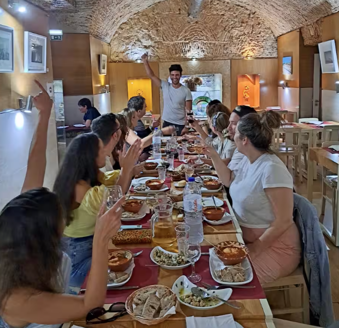 Group of people enjoying a meal together in a restaurant with arched brick ceiling, some are raising hands, and a man standing at the head of the table smiling.