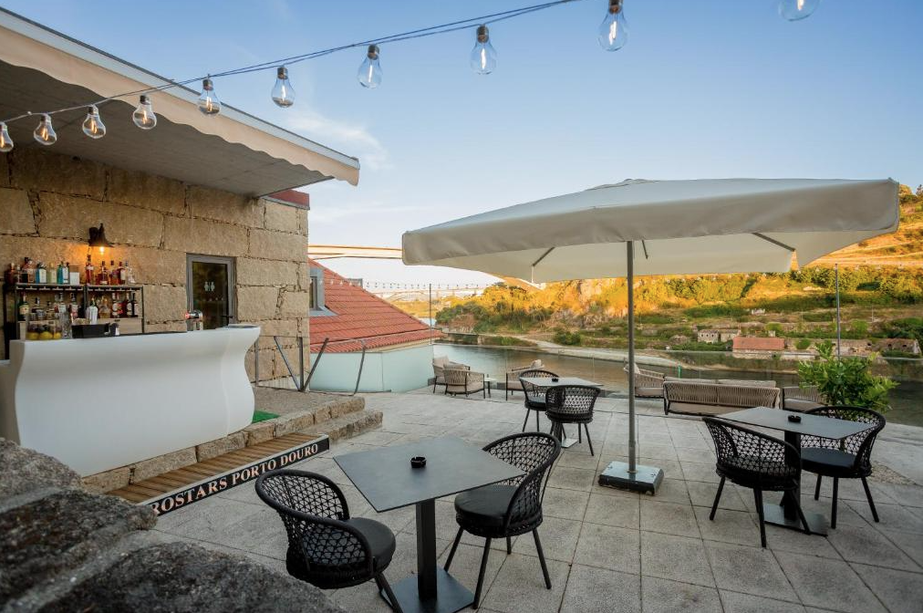 Outdoor terrace with tables, chairs, a large umbrella, string lights, a bar area, and a scenic river view with rolling hills.