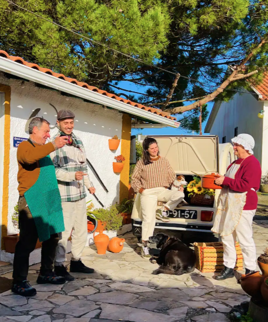 A group of five people gathered outdoors near a house, celebrating with drinks. One woman is holding a cake, and a black dog is lying on the ground nearby. There are pumpkins and potted plants as decorations, with a tree in the background under a cle