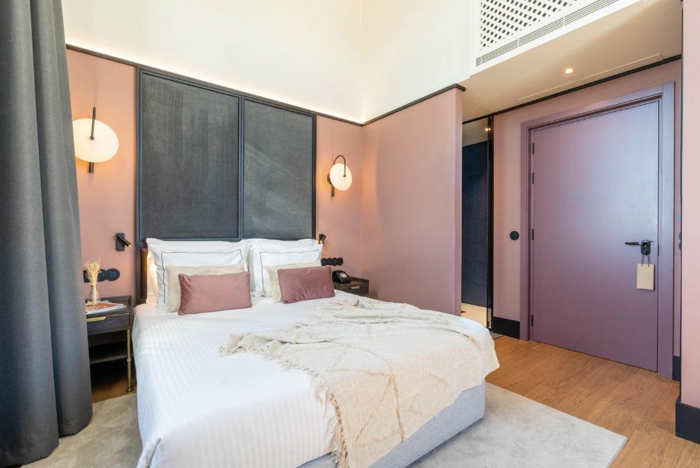 A modern bedroom with a neatly made bed, pink walls, wall-mounted lamps, and a purple door.