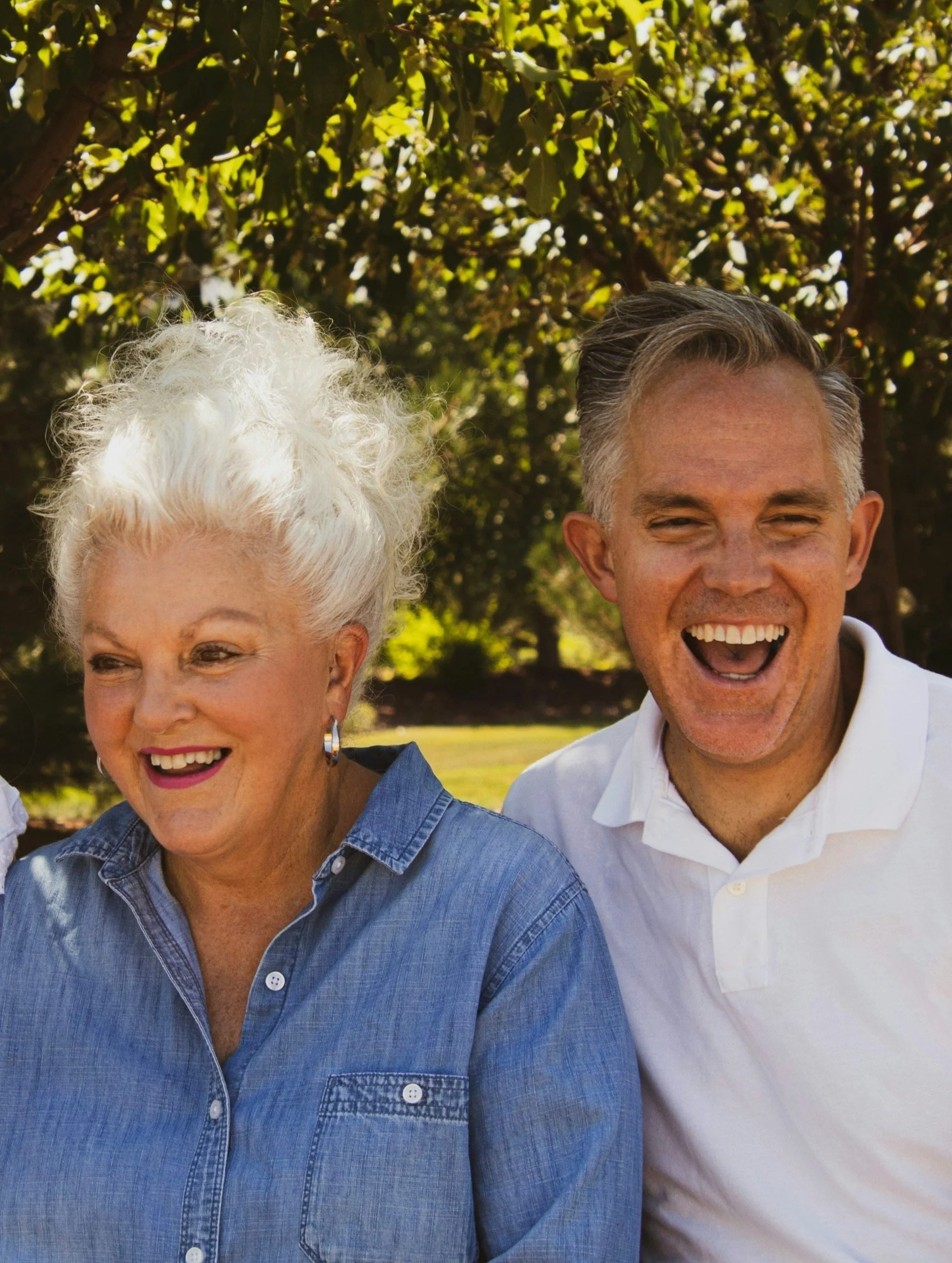 Smiling older woman in a denim shirt and a happy man in a white shirt standing outdoors under trees.