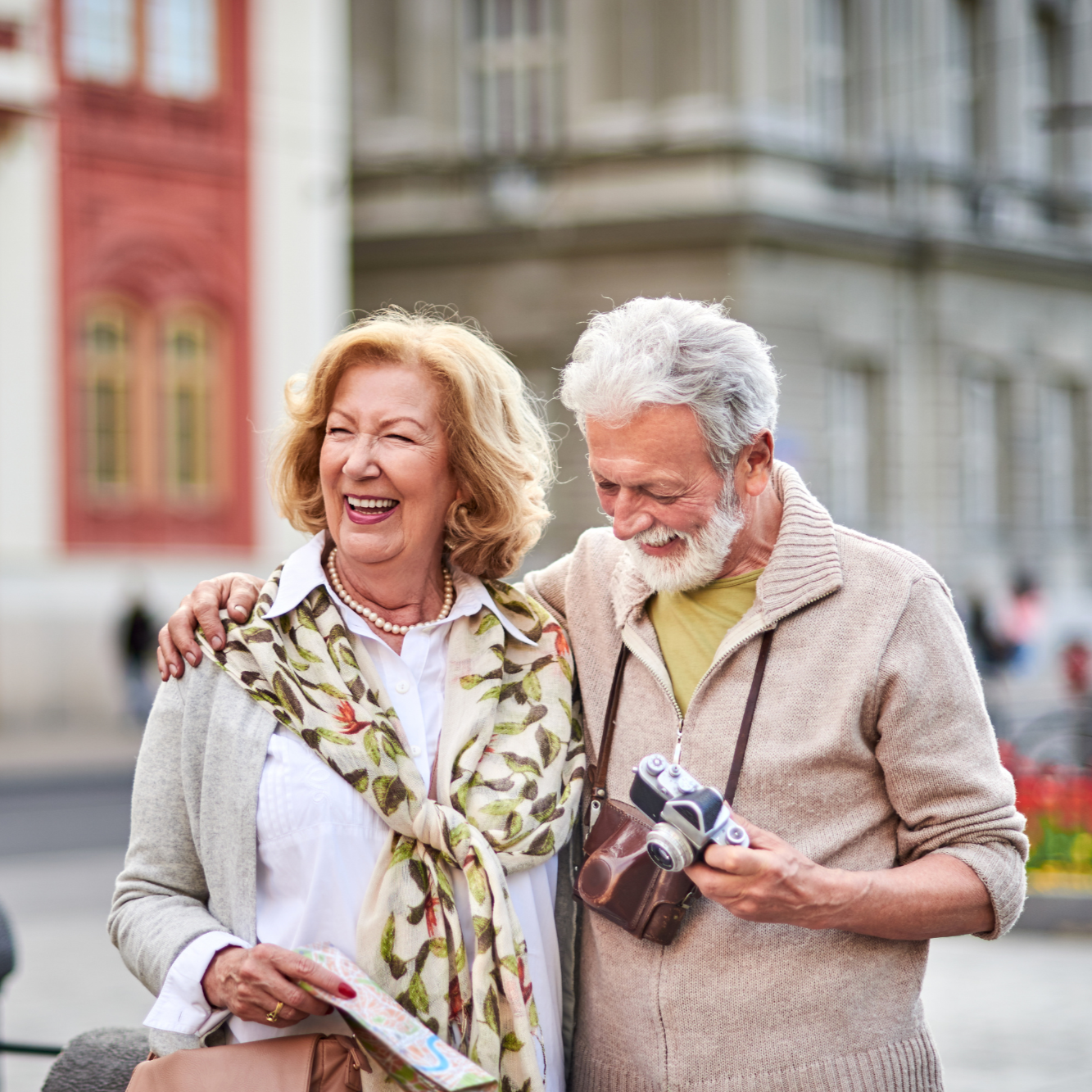 An elderly couple, smiling and laughing together outdoors in an urban setting, with the man holding a vintage film camera.