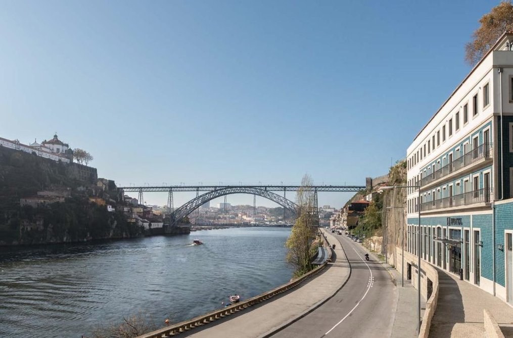 A river with a bridge crossing it, flanked by buildings and hills, under clear blue sky.