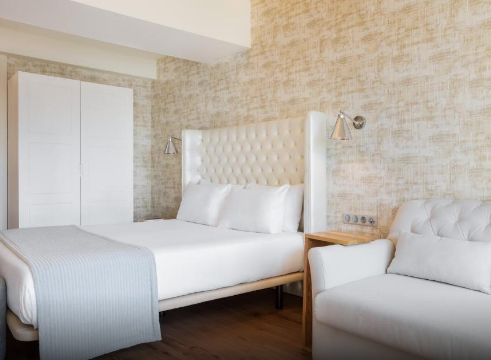A bedroom with a white queen bed, tufted headboard, white pillows, a white comforter, a wooden side table, a white sofa, and a textured beige wall.