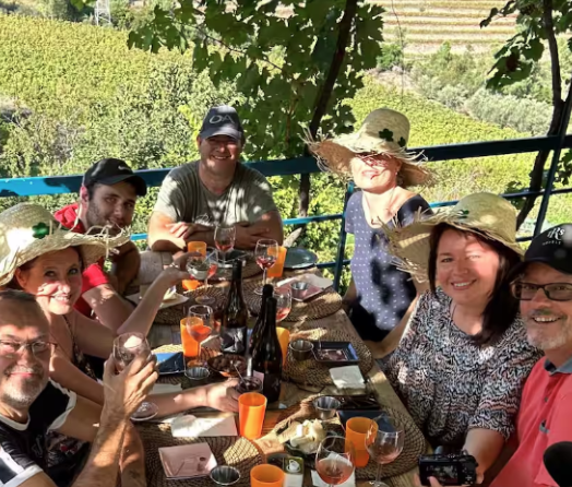 Group of people sitting at a table outdoors, smiling, with drinks and bottles, under a canopy of trees.