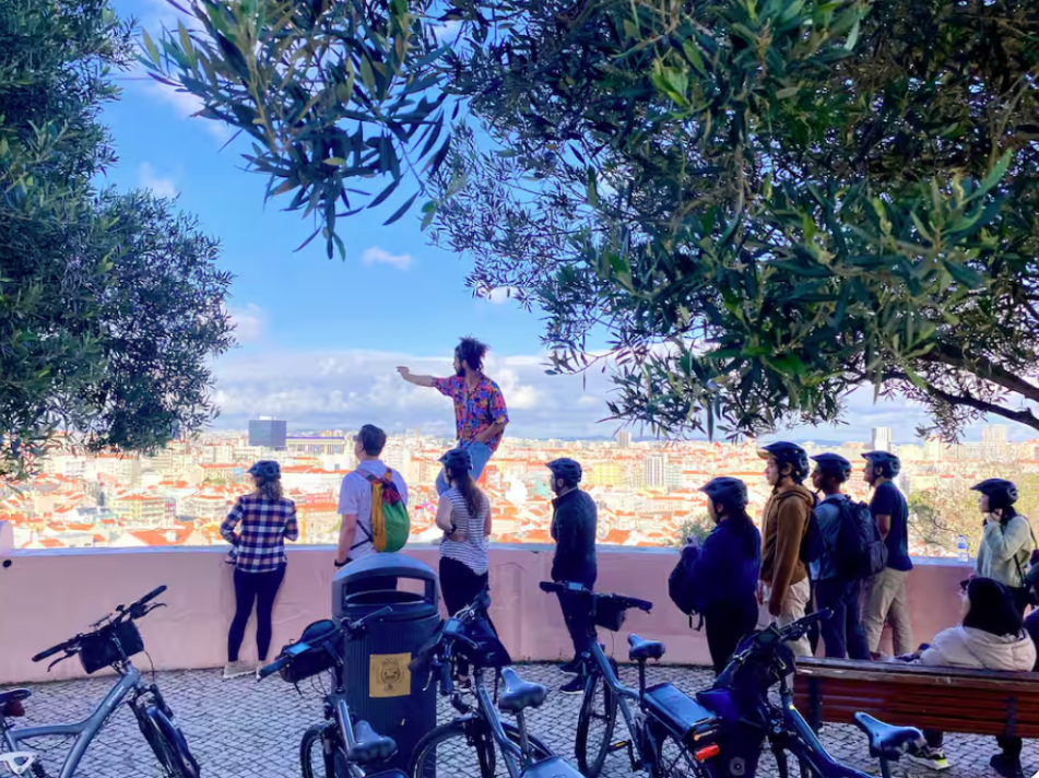 A group of people with helmets, standing on a lookout point, looking at a cityscape with a person standing on a wall, gesturing towards the view, surrounded by trees and bicycles.