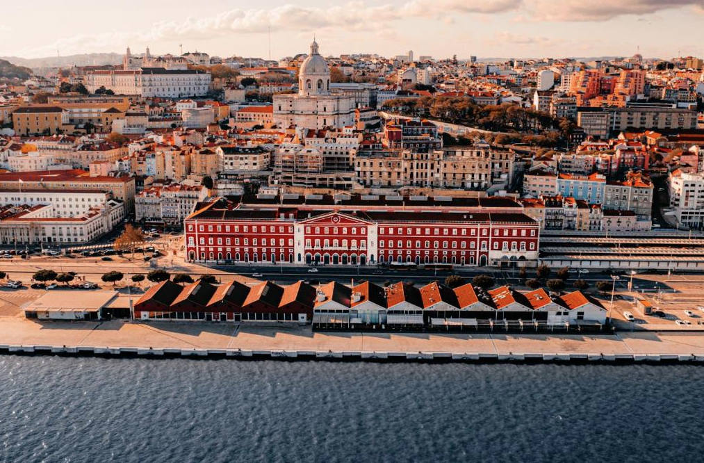 Aerial view of a cityscape showing a waterfront with red-roofed buildings, a large red and white historic building, and a skyline with various buildings and monuments in the distance.