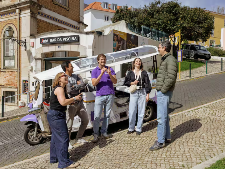 Five people standing and chatting next to a small purple and white shuttle bus on a cobblestone street in a European town.