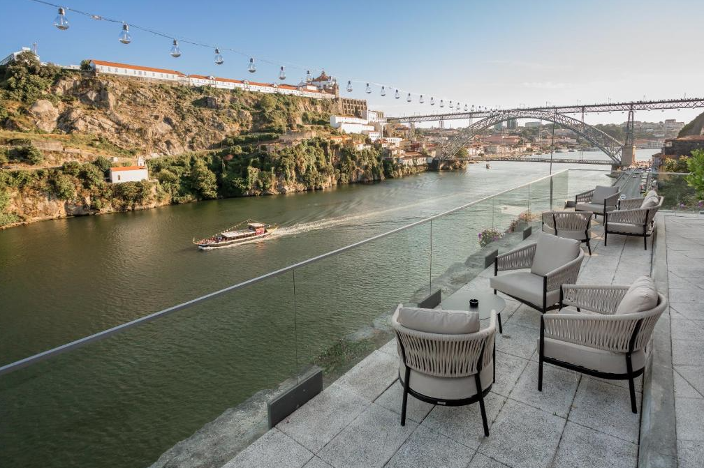 Outdoor balcony with white cushioned chairs and small tables overlooking a river with a boat, hillside buildings, and a bridge in the background.