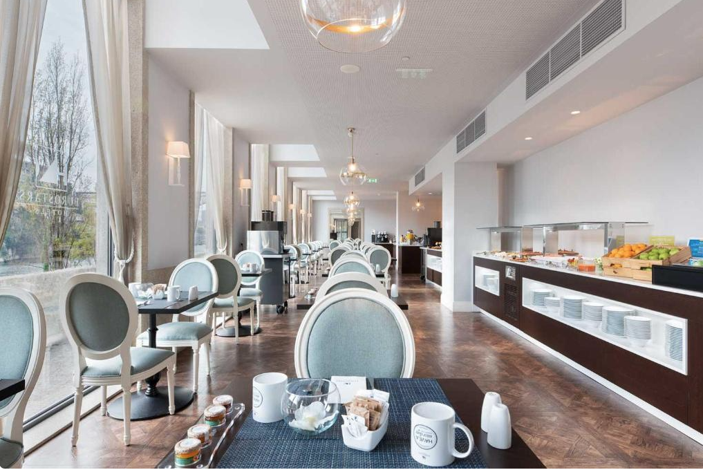 A dining area with tables and chairs, set with mugs, sugar, and condiments. A buffet with fruits and breakfast items is along the right wall. Large windows offer outside views.