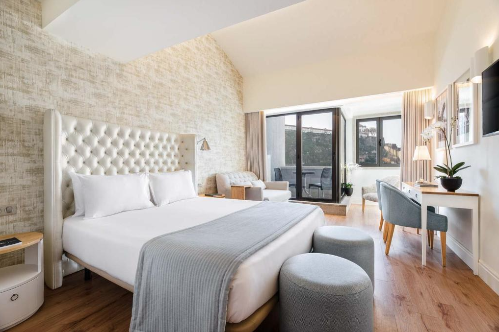 A bright hotel room with a large white bed, a tufted headboard, side tables, seating area with chairs, a desk, and a balcony door leading outside.