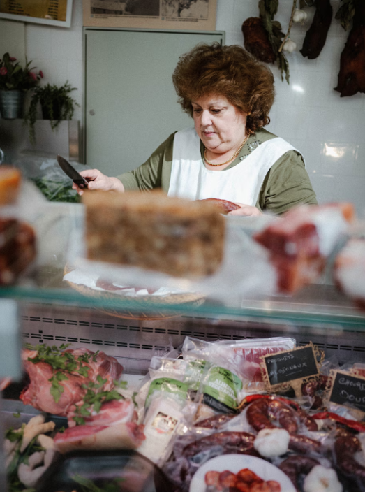 A woman with curly hair examining meat in a deli or butcher shop, looking at her phone, surrounded by various cuts of meat and sausages.