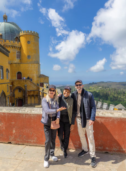 Three people standing in front of a colorful castle with yellow walls and a dome, with a scenic landscape and blue sky in the background.
