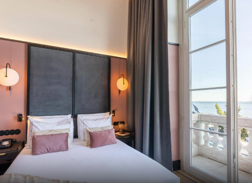 A modern hotel bedroom with a large bed, pink pillows, and a view of the ocean through glass doors leading to a balcony.