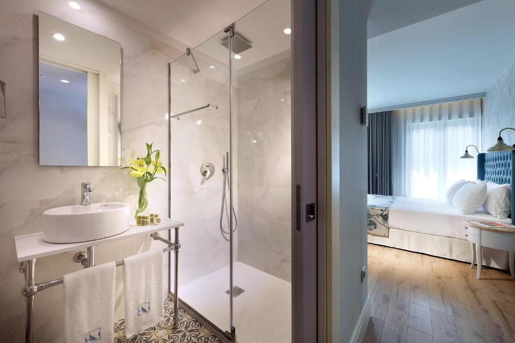 Modern hotel bathroom with a glass shower, white sink with a mirror, yellow flowers, and towels, next to a bedroom with a white bed, blue headboard, and natural light.
