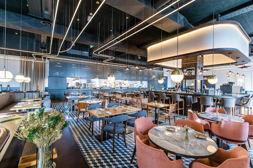 A modern, upscale restaurant with various table arrangements, stylish chairs, a bar area with barstools, decorative lighting fixtures, and a patterned floor.