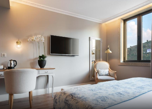 Modern bedroom with a bed, a chair, a desk with a kettle and flowerpot, a wall-mounted TV, a display cabinet, and a large window showing mountains and blue sky.