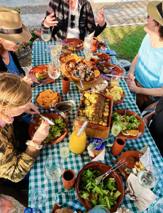 A group of people having a meal outdoors at a table with various dishes, drinks, and a checkered tablecloth.