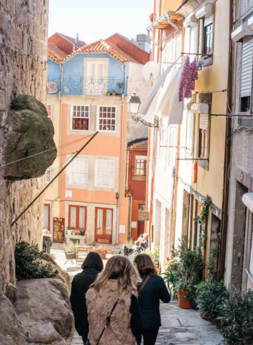 People walking down a narrow, colorful street lined with flowers and laundry hanging out to dry in a European city.