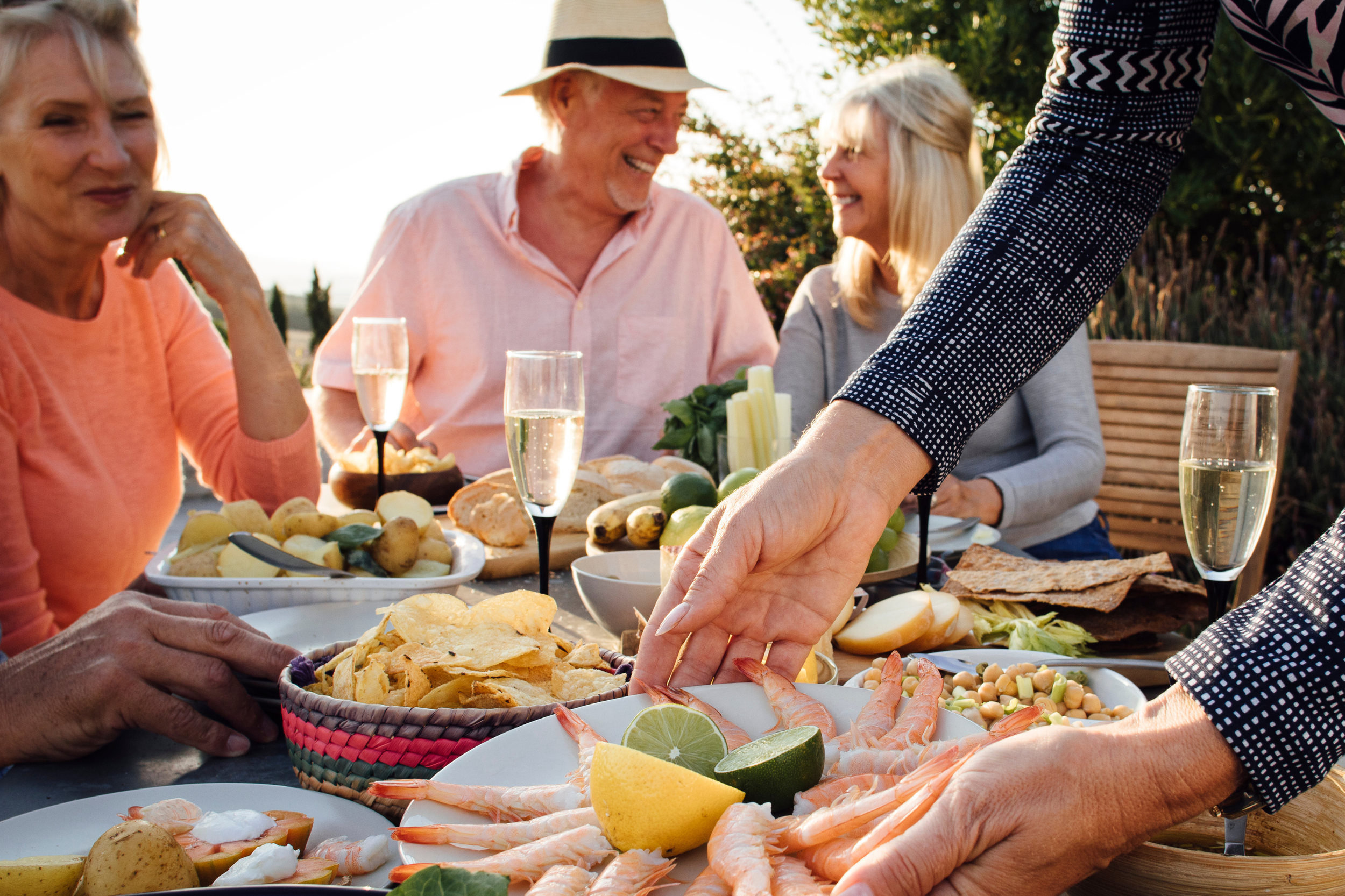 Group of adults enjoying an outdoor seafood meal, with shrimp, lemon, lime, chips, potatoes, and drinks on the table.
