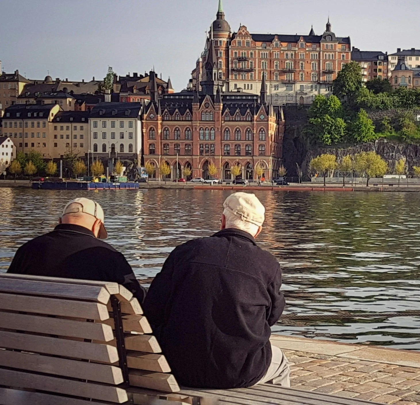 Two people sit on a bench by a waterfront, with historic, ornate buildings visible across the water in the background.