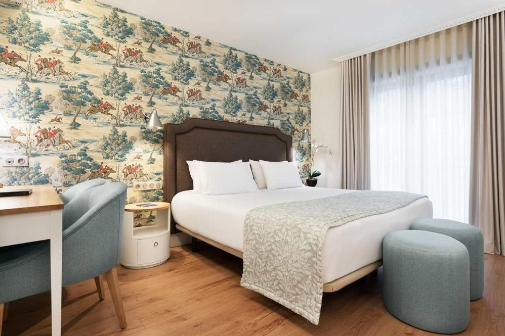 Hotel room with a bed, blue upholstered chairs, a wooden desk, and a tapestry wallpaper featuring trees and horses.