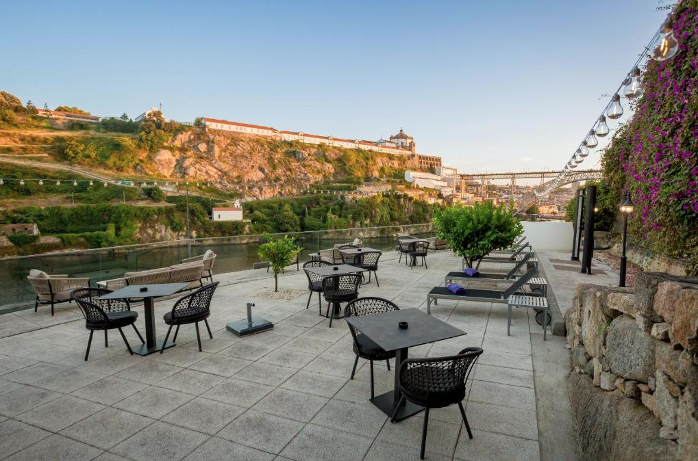 Outdoor terrace with tables, chairs, and lounge chairs overlooking a river and hillside with buildings and a bridge in the background, decorated with string lights and purple flowers.