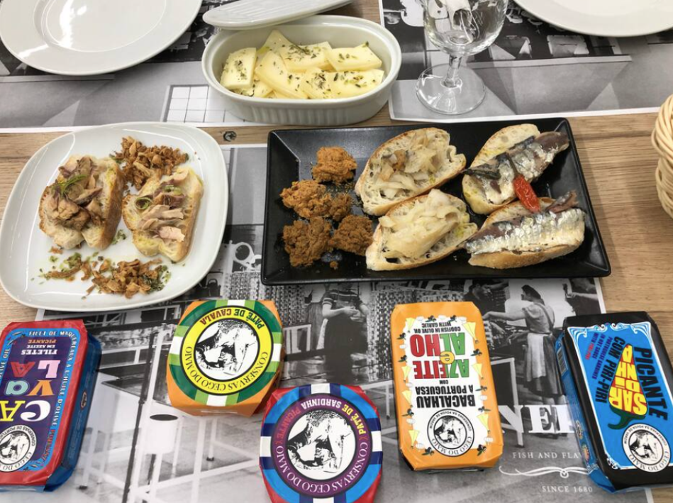 An assortment of Italian cuisine including breaded fried fish, cheese, and cured meats on a black platter, a white dish with bread topped with sliced ham and green onions, and several packages of La Vache Quirit cheese, on a table set with glasses an