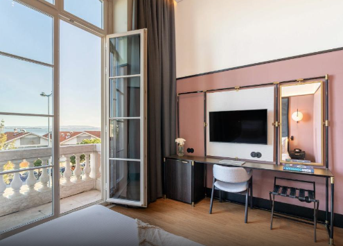 Hotel room with a balcony, a wall-mounted TV, a desk with a chair, and a large window allowing natural light