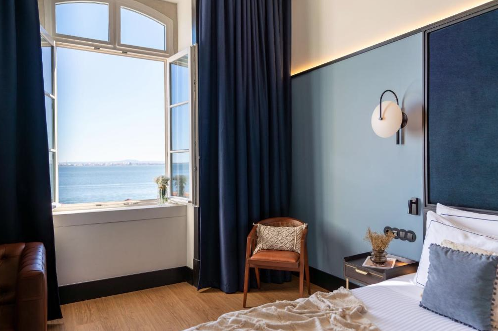 A bedroom with a large open window showing a body of water and a city skyline in the distance. The room has a blue accent wall, dark curtains, a modern bedside lamp, a small side table with a decorative plant, and a portion of a bed with white beddin