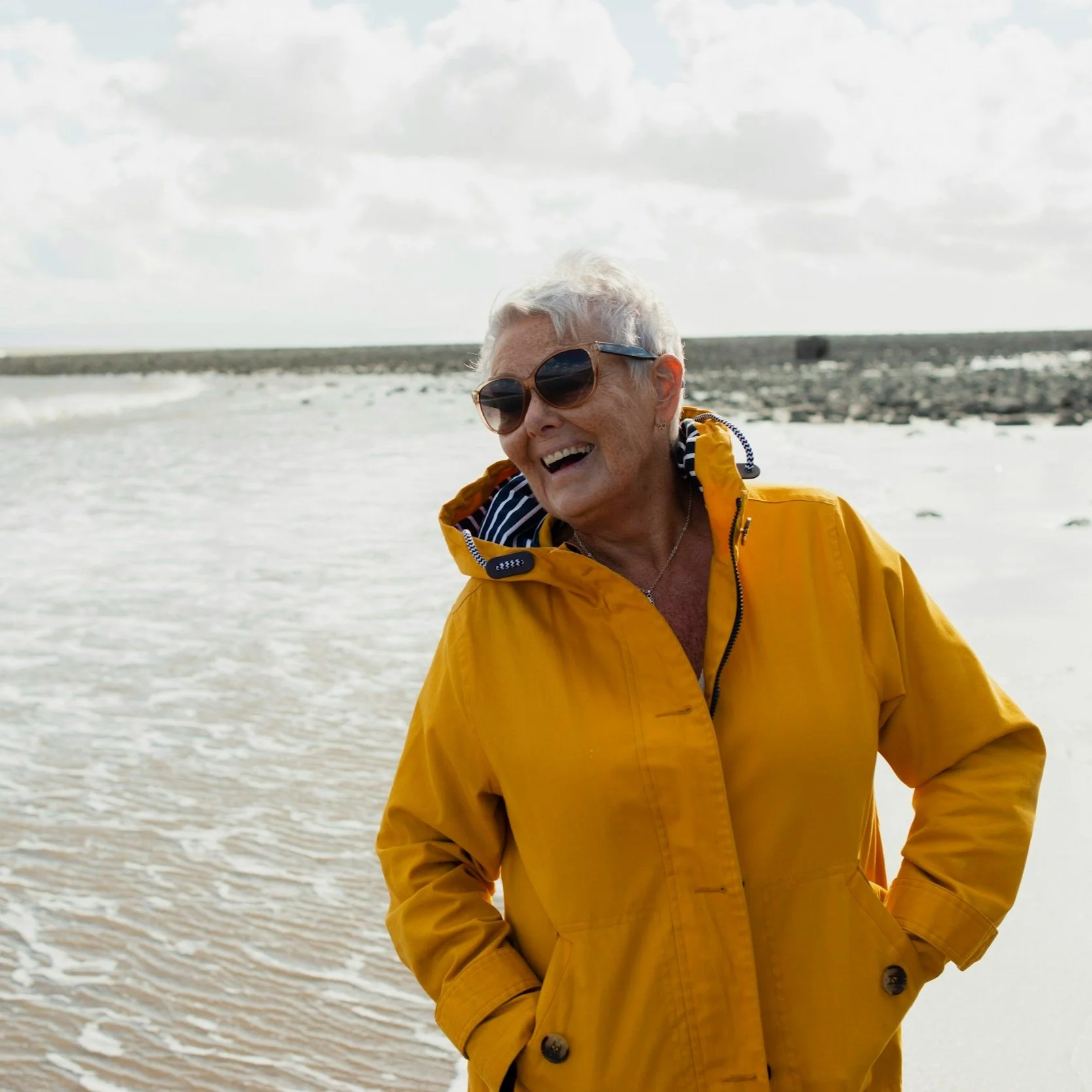 Elderly person wearing a yellow jacket and sunglasses standing on a beach with the ocean and sky in the background.