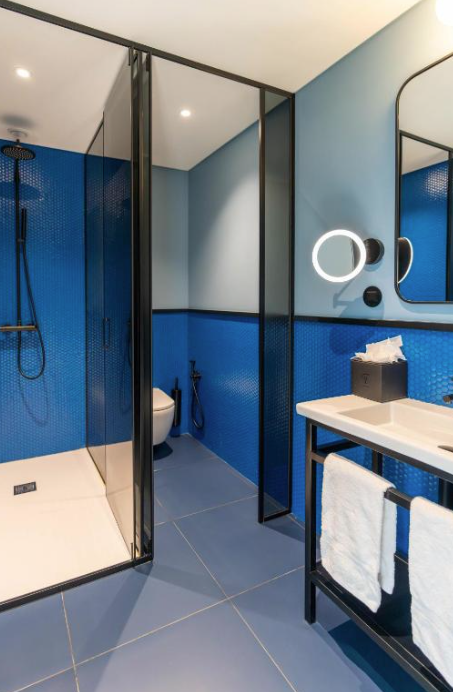 Modern bathroom with blue tiled walls, a glass shower enclosure, a wall-mounted toilet, a vanity with a white sink, a mirror, and a circular illuminated mirror. Towel hanging on a black metal rack and a tissue box on the vanity.