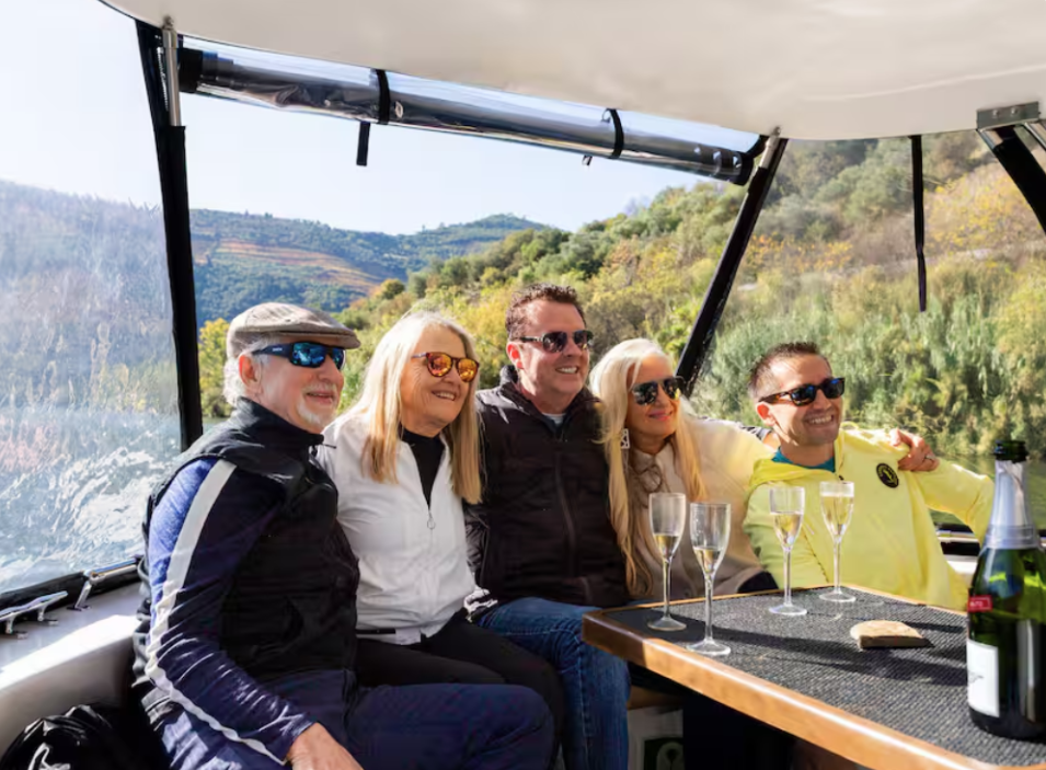 Group of five people sitting on a boat enjoying a scenic river cruise with greenery and hills in the background, some holding champagne glasses.