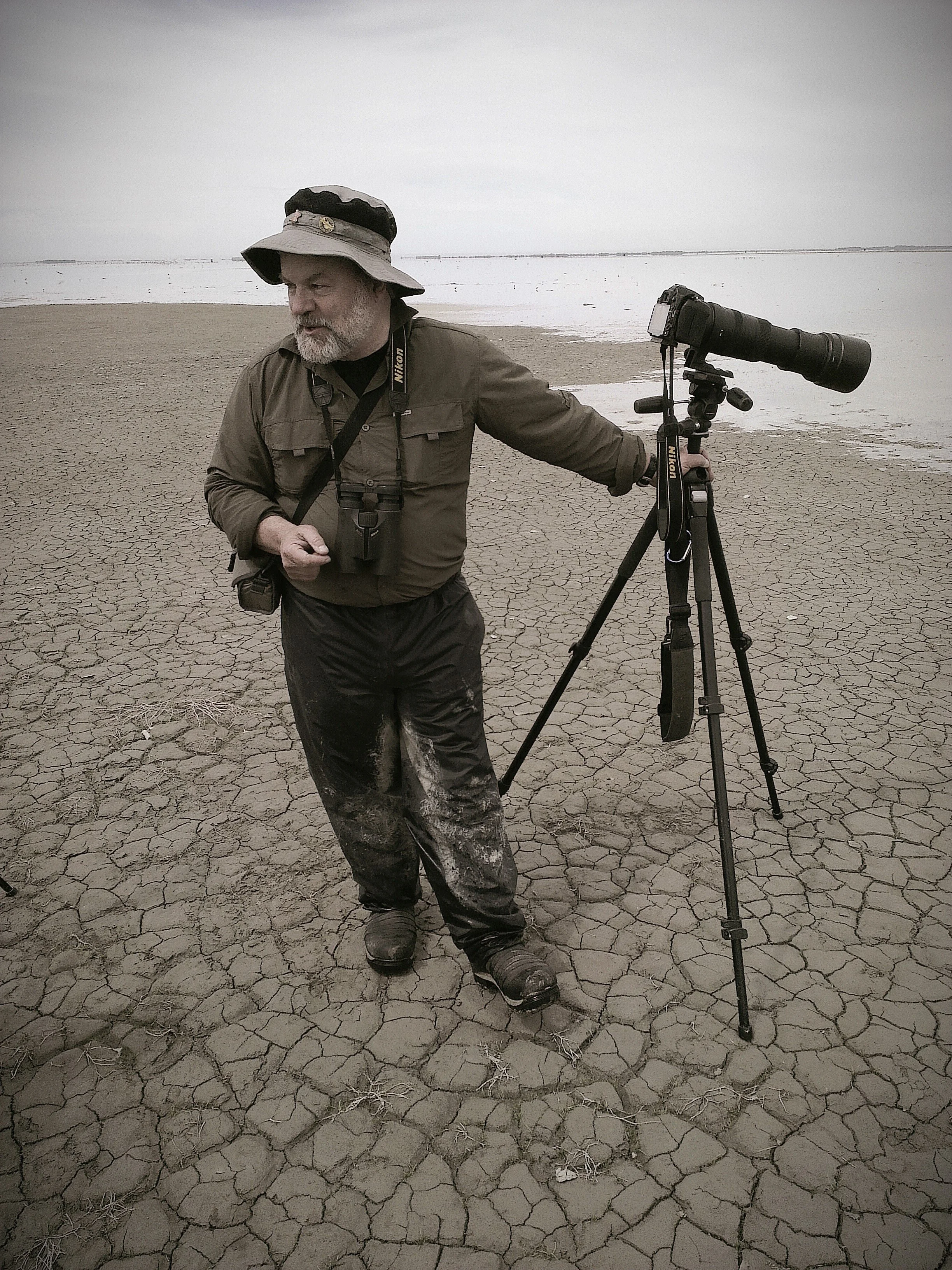 A man outdoors in a dry, cracked landscape with a body of water in the background, wearing a wide-brimmed hat, waterproof clothing, and holding a camera with a long lens on a tripod.