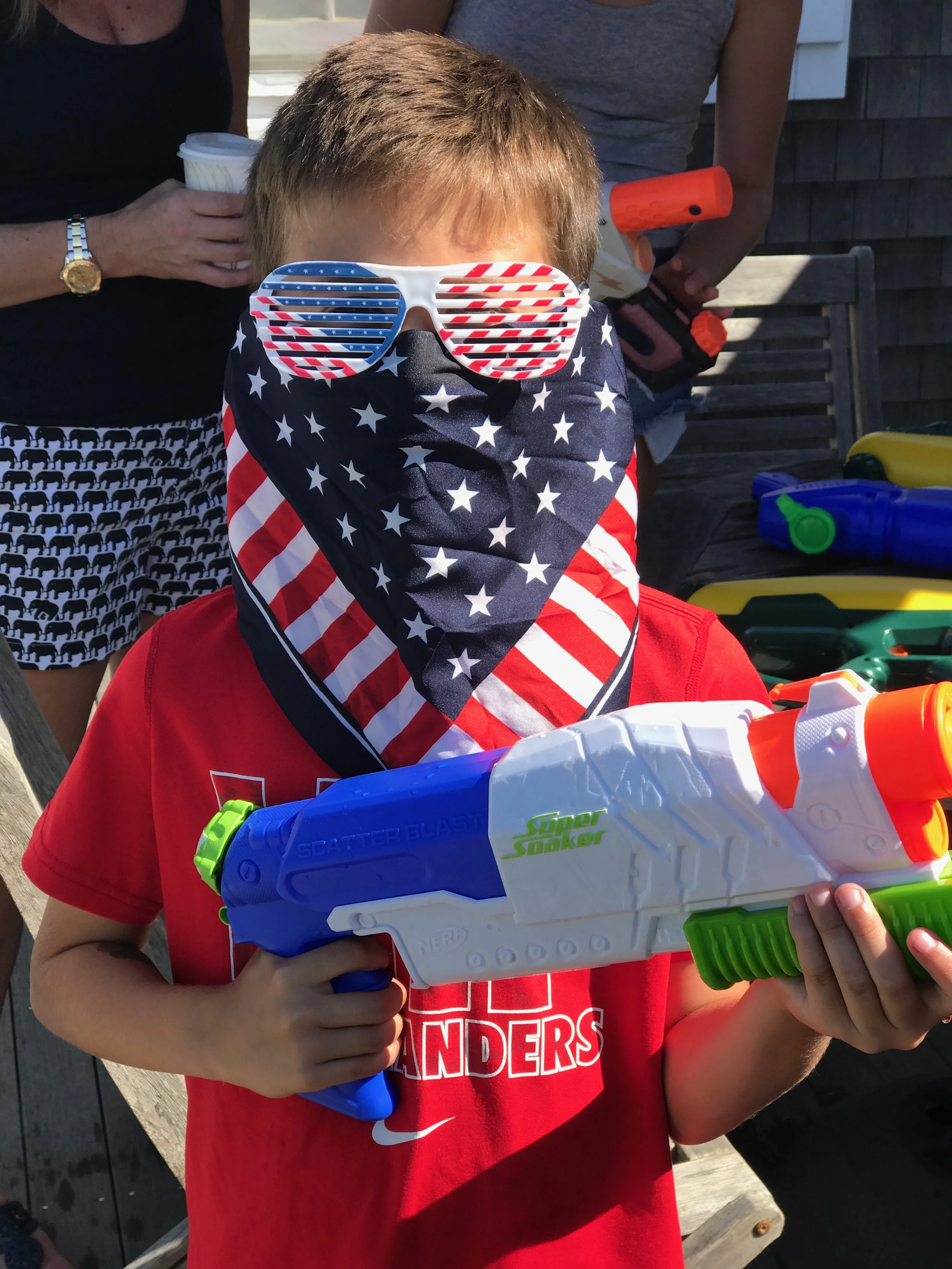 A child wearing sunglasses with American flag design, a patriotic bandana covering his face, and a red shirt. He is holding a toy water gun.
