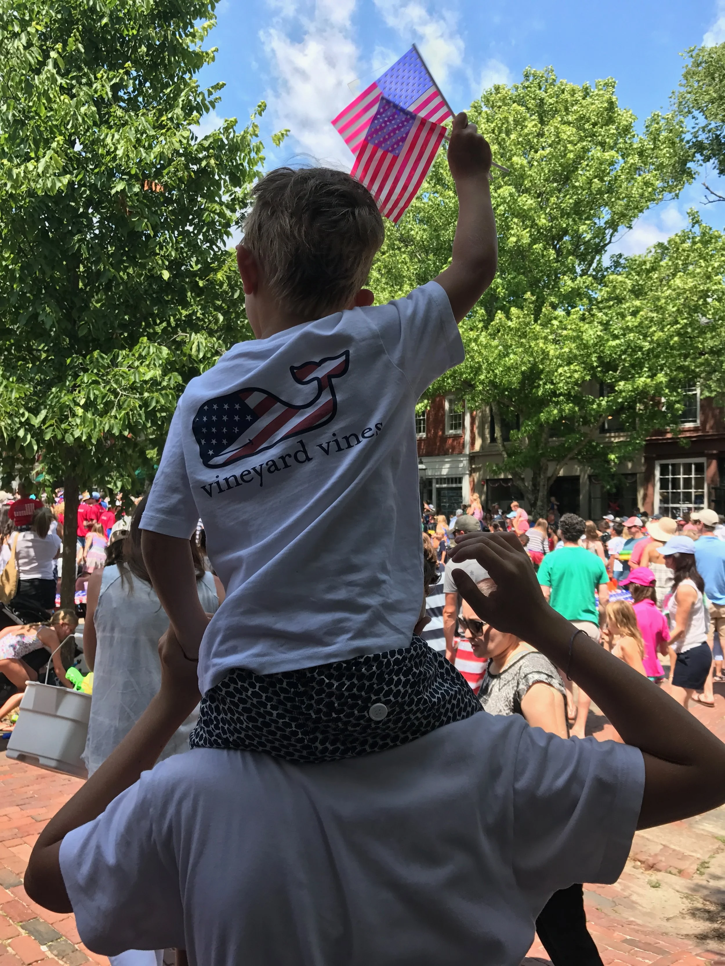 A young boy sitting on someone's shoulders, waving American flags during a crowded outdoor celebration. The boy is wearing a white t-shirt with a logo and text, and the background shows numerous people dressed in patriotic colors, with trees and buil