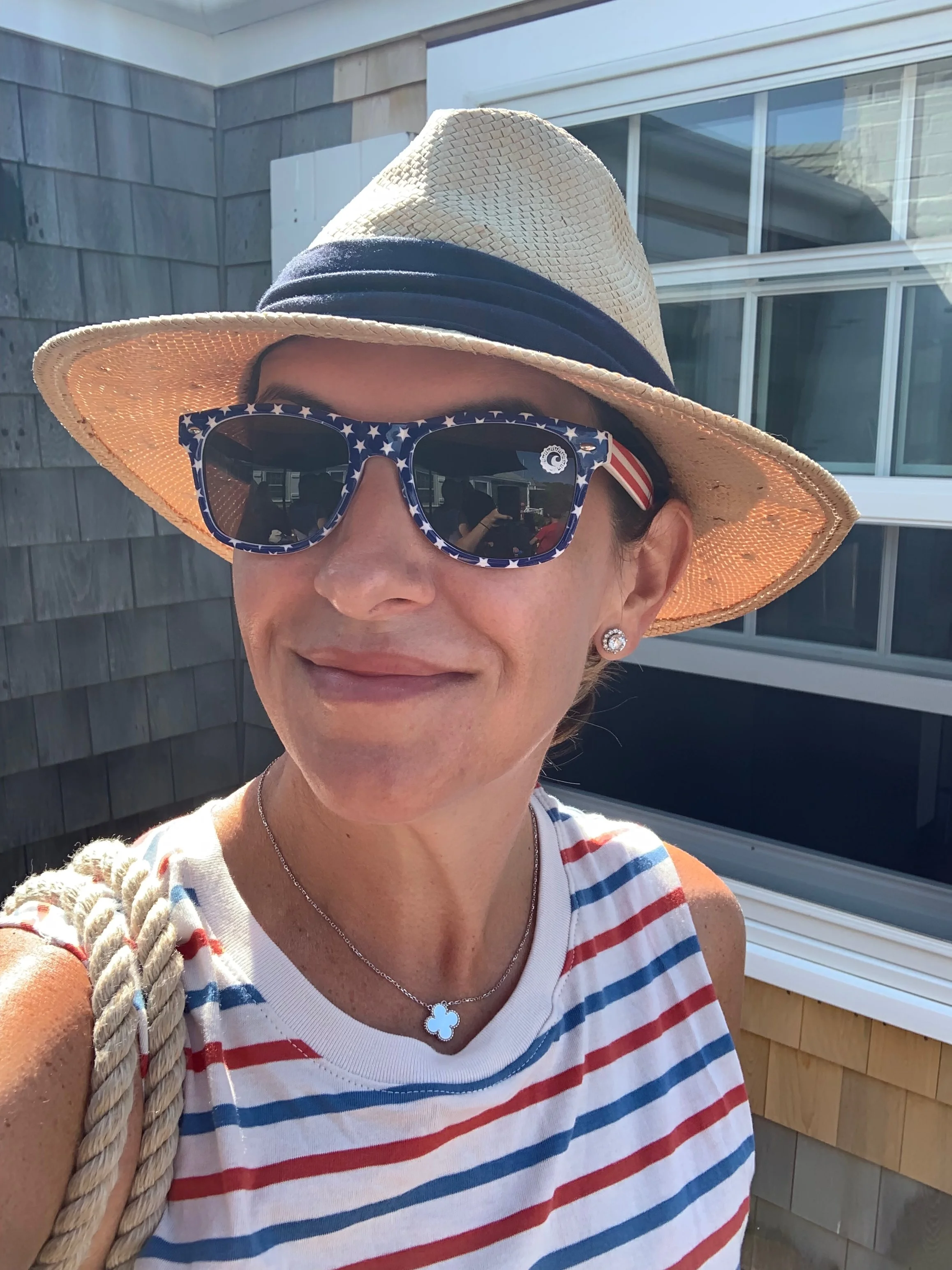 Woman wearing a straw hat, American flag-themed sunglasses, and a striped sleeveless top, standing outdoors in front of a building with large windows and grey siding.