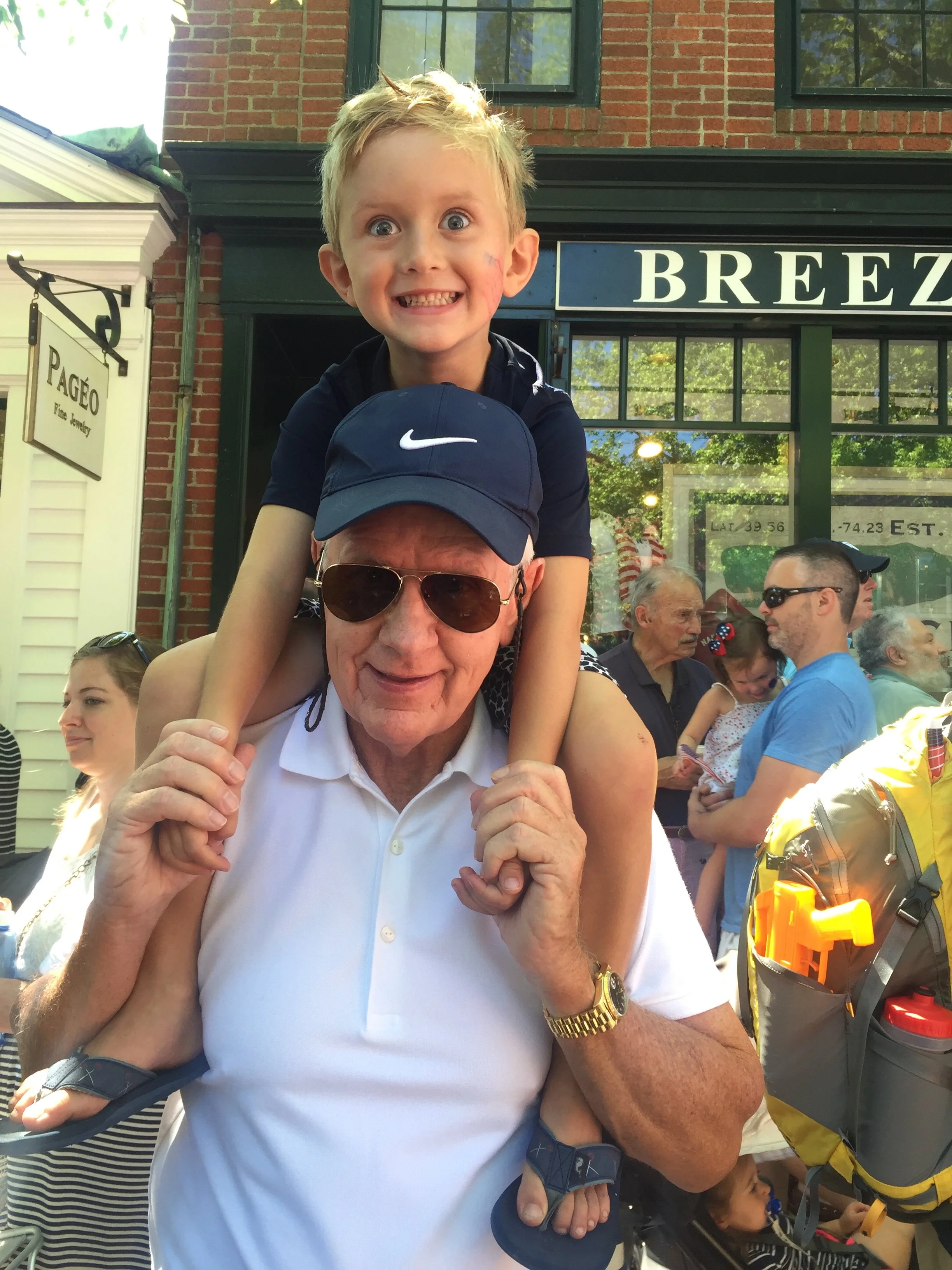 An elderly man wearing sunglasses and a white polo shirt is carrying a young boy on his shoulders. The boy has blonde hair, blue eyes, and is smiling broadly. The background shows a crowd of people gathered outdoors in front of storefronts, with some