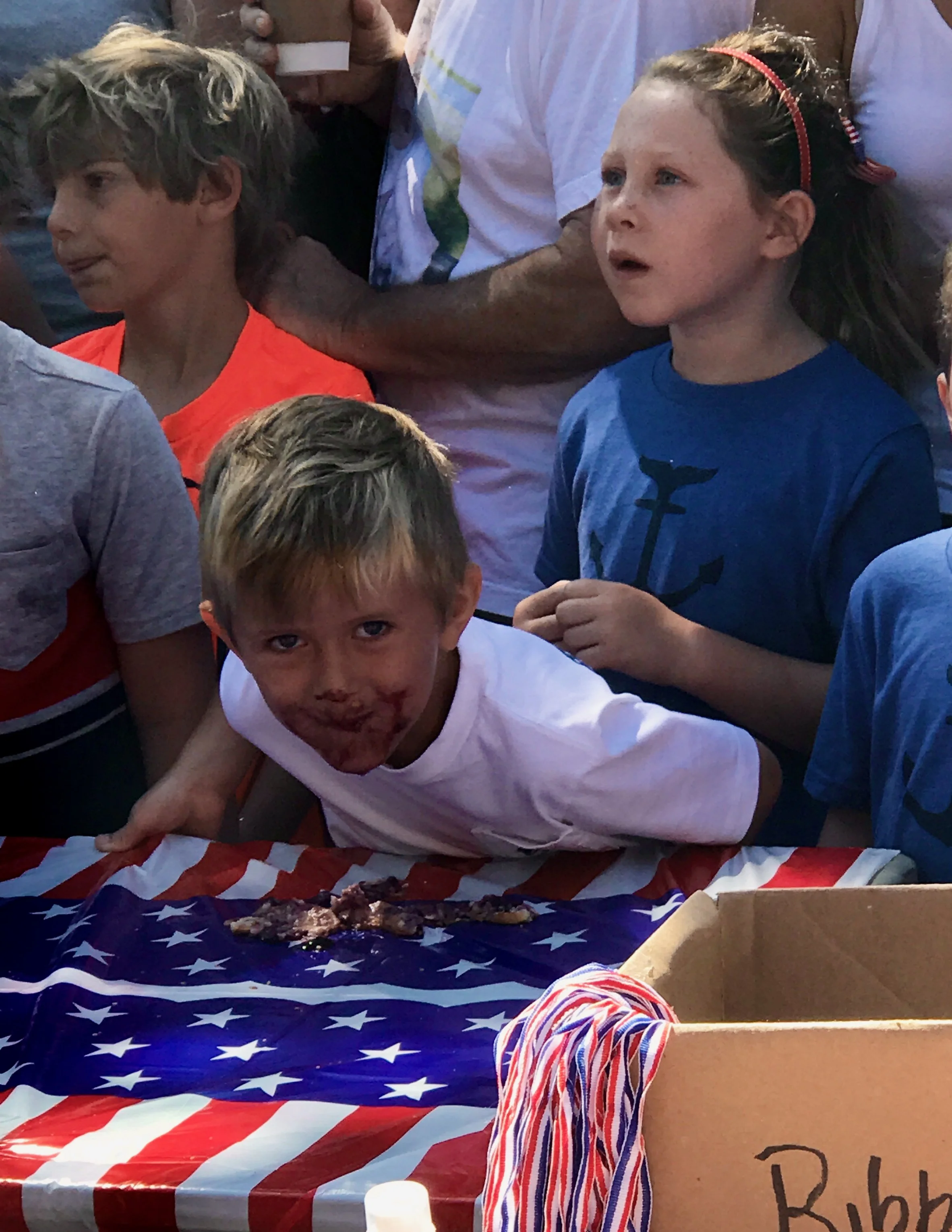 Children gathered around a table with an American flag tablecloth, one boy with a dirty face and a big smile, another girl looking surprised, and others partially visible. There is a box with red, white, and blue striped string nearby.