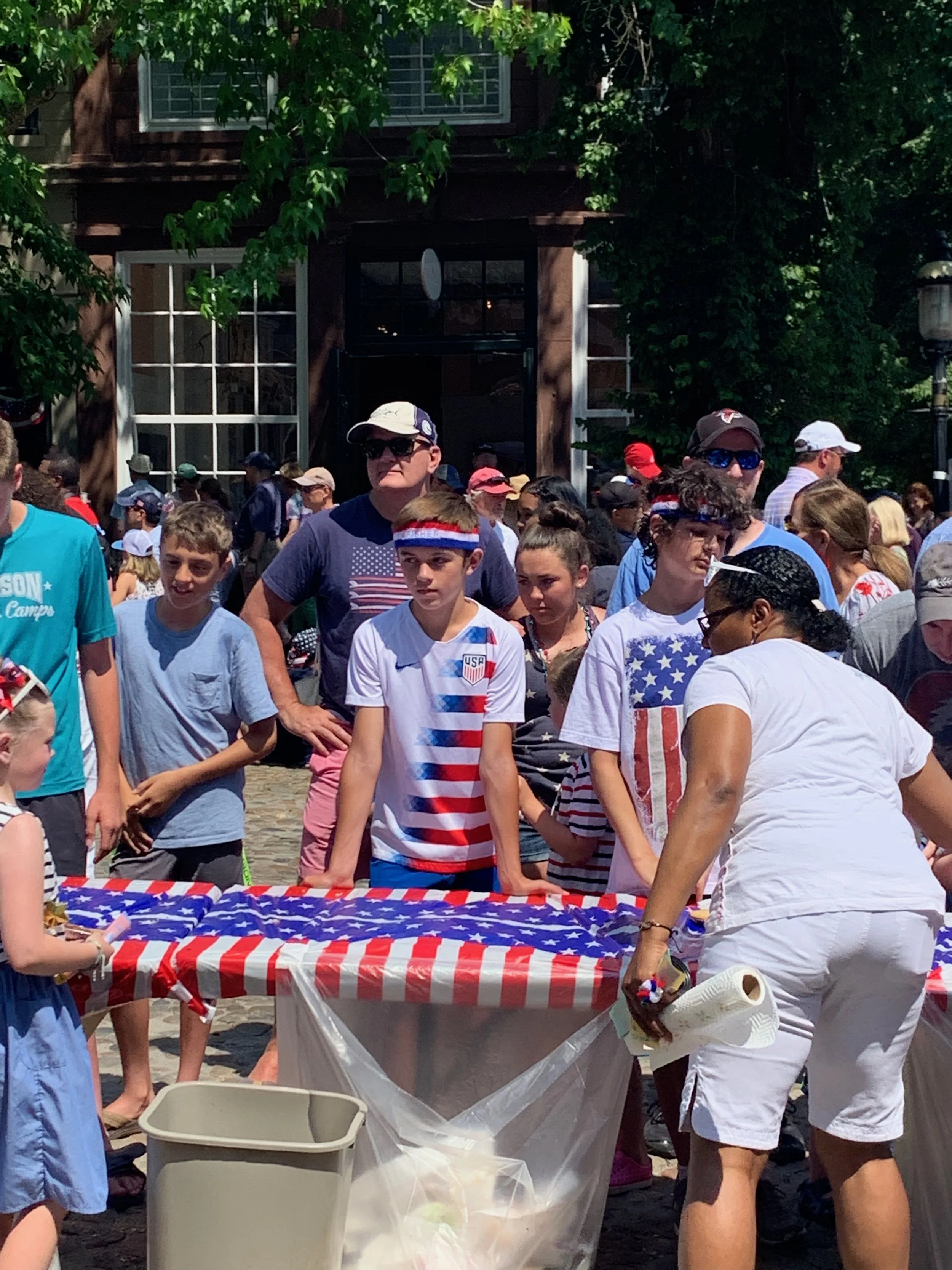 Crowd of people gather outdoors around a table decorated with an American flag-themed tablecloth, with many wearing patriotic clothing and accessories.