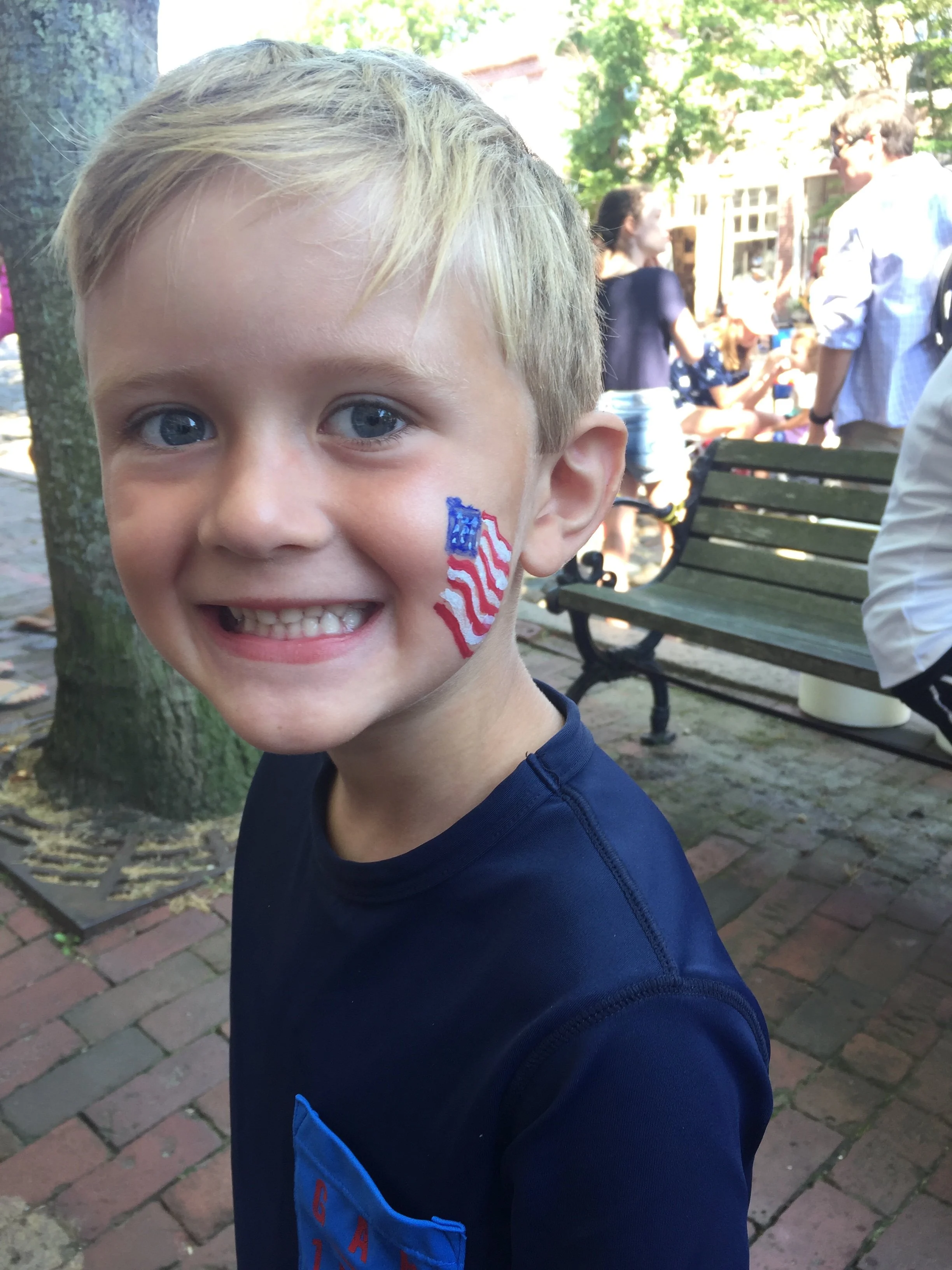 A young boy with blond hair and blue eyes smiling, with an American flag face paint on his cheek, outdoors in a park or street setting with people in the background.